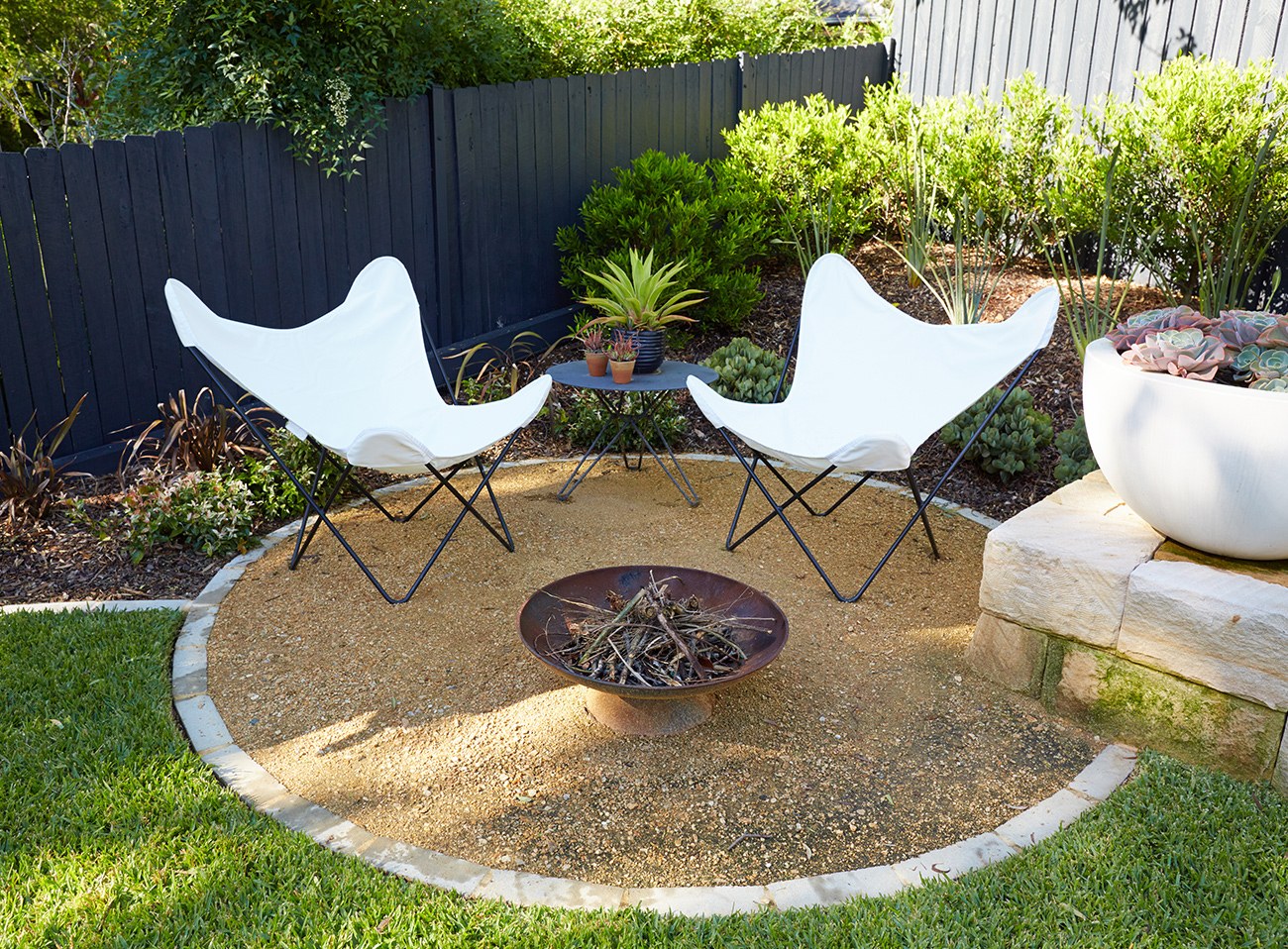 Landscape design and construction with a circular gravel fire pit terrace, stone edging, white sling chairs and layered planting against a dark fence.