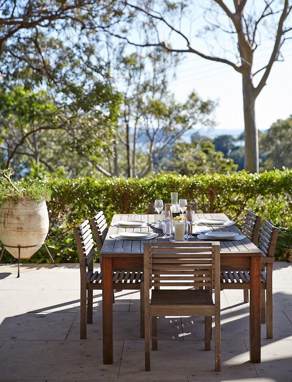 Landscape design and construction with a paved outdoor dining terrace, timber table setting, clipped hedge screen and treed water view beyond.